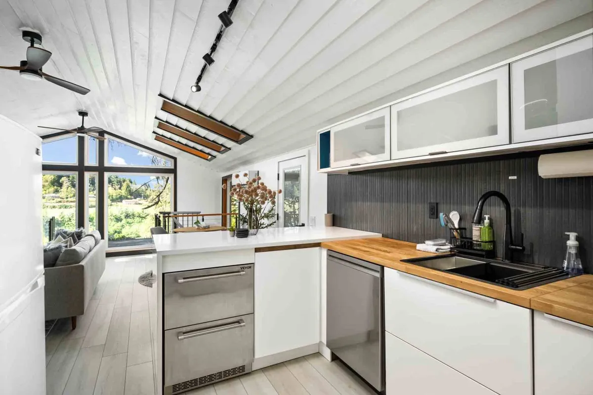A stylish, contemporary kitchen with a spacious island, modern appliances (including a built-in oven, stove, and dishwasher), and decorative touches like a flower vase. Large windows provide abundant natural light and offer a scenic backdrop while prepping or enjoying meals.