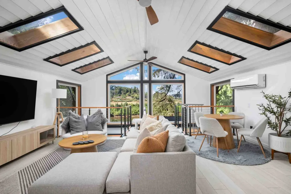 A welcoming open-plan living and dining space with large windows that reveal a peaceful view of the natural landscape. The area includes a cozy sofa with colorful cushions and a stylish dining table, illustrating the seamless flow between lounging and dining environments.
