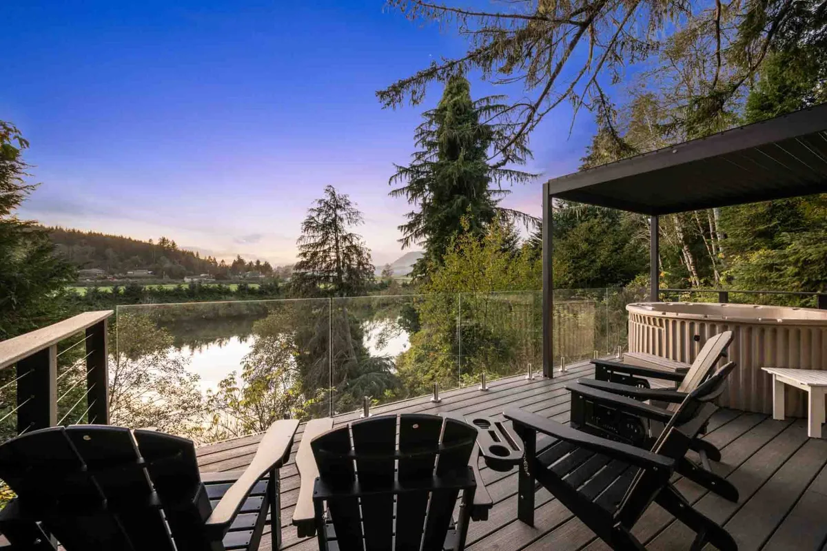 This exterior image captures a serene riverside setting with Adirondack chairs arranged near a hot tub, all positioned to look out over the Nehalem River and surrounding trees. The relaxing seating area and bubbling hot tub are bathed in soft, natural light, evoking the peacefulness of riverside living and the promise of tranquil mornings or evenings.