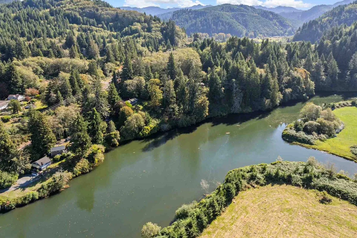 A sweeping view of the Nehalem River winding through a dense forest, with a distant meadow and mountain range. The wide-open sky suggests opportunities for stargazing and conveys the peacefulness and natural beauty of the location.