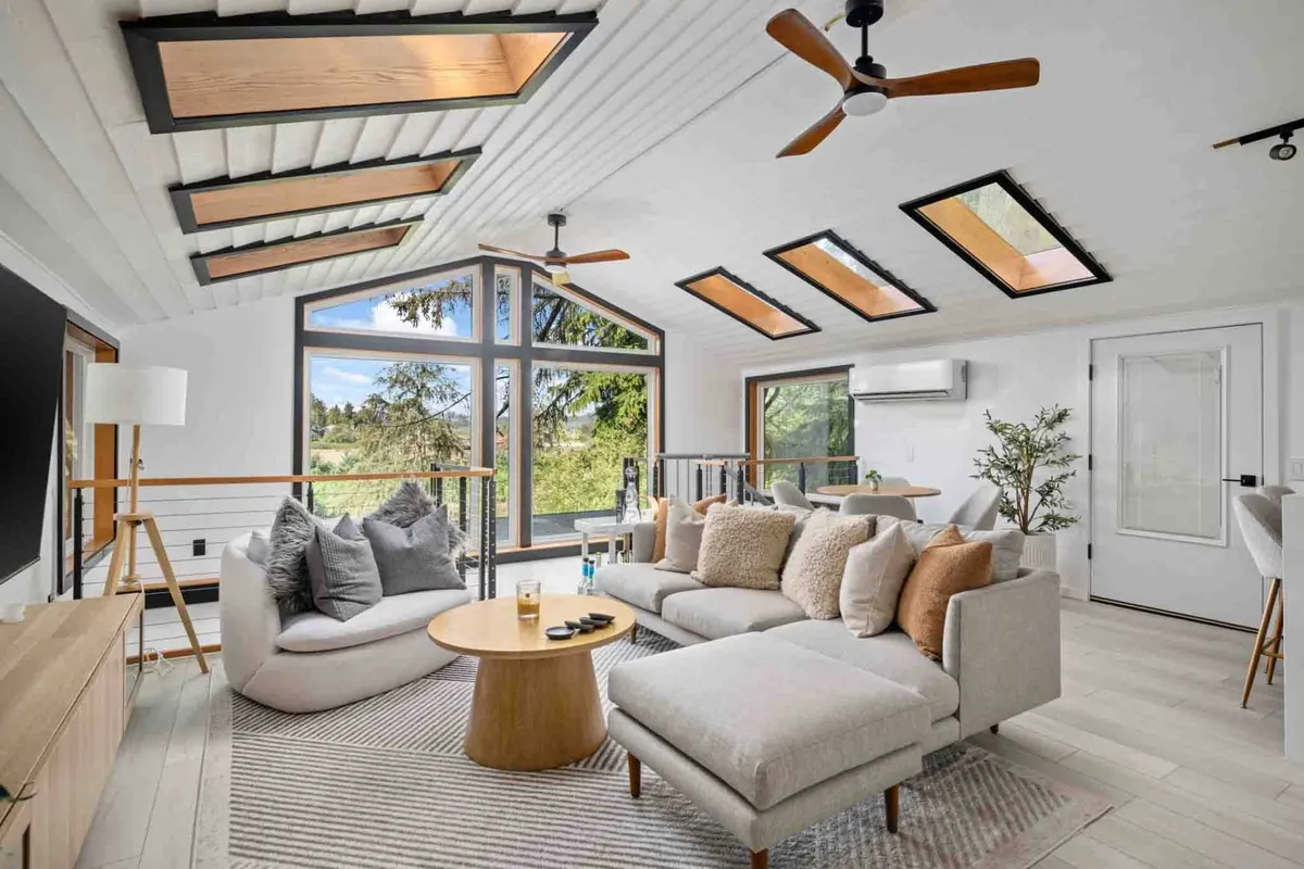 A sunlit living area featuring expansive windows that frame the riverside scenery, plush and comfortable seating, a wooden ceiling with skylights that flood the room with natural light, and a seamless blend of modern and cozy design. The setting is ideal for relaxation, social gatherings, and enjoying the tranquil landscape outdoors from the comfort of indoors.