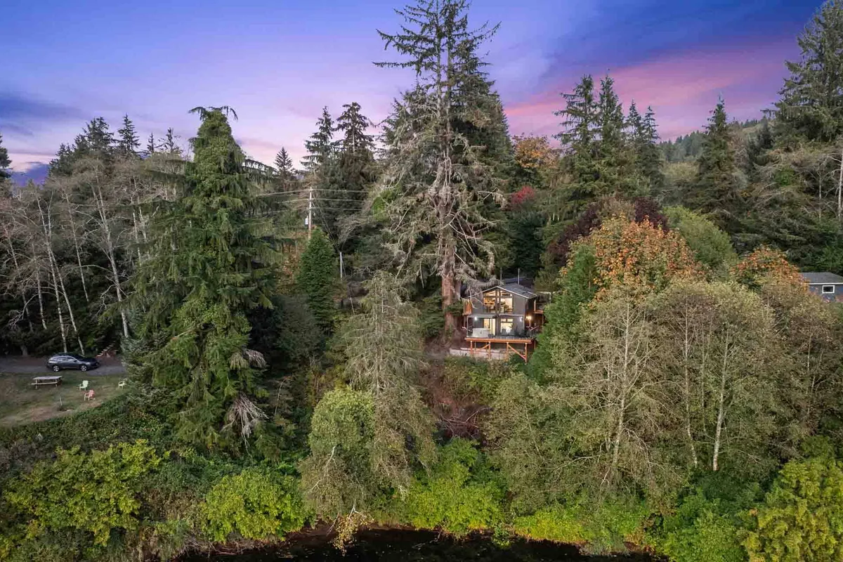 The exterior of the house with its clean lines overlooks a tranquil river, framed by tall trees and natural landscaping. The setting suggests immediate access to nature for riverside strolls, relaxation, or wildlife watching.