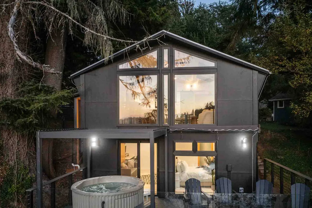 A cozy outdoor scene featuring a large hot tub nestled by the house, with expansive glass windows reflecting the surrounding trees. An inviting seating area sits nearby, all set within lush greenery, creating a peaceful retreat for relaxation and stargazing.