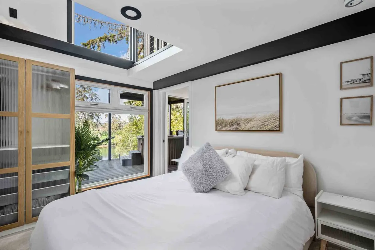 A serene bedroom space featuring a cozy bed with soft bedding, large windows that let in natural light, subtle artwork above the headboard, and a touch of greenery from a potted plant. The room feels welcoming, peaceful, and offers scenic outdoor views for relaxation.