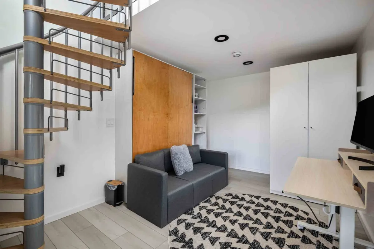 A cozy, multi-functional living space featuring a spiral staircase, sofa, rug, and a desk with a TV. The area balances comfort, relaxation, and productivity, with a workspace element visible within a social setting.