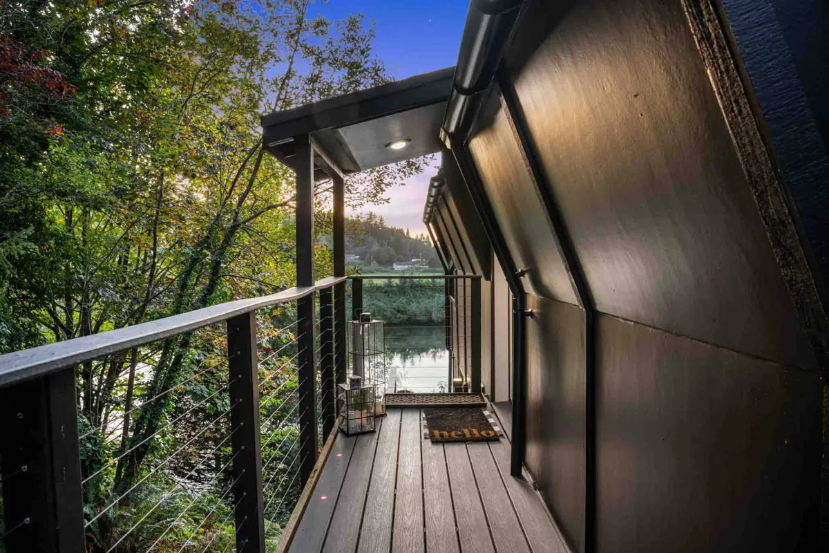 A tranquil wooden deck overlooking the river, featuring a decorative lantern and a cozy welcome mat. The deck is designed for enjoying the outdoors with comfortable seating and a direct view of serene water and lush greenery, ideal for sipping morning coffee or relaxing at sunset.