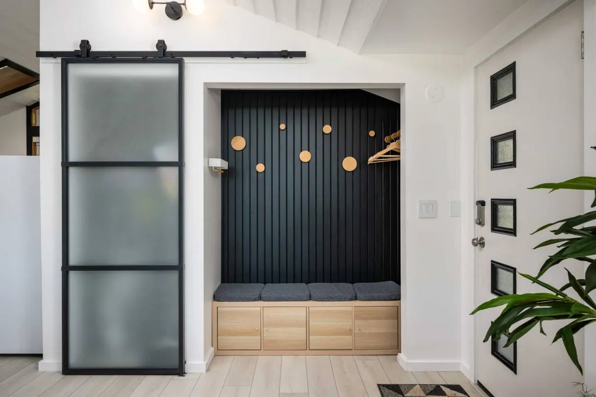 An inviting interior entryway with a sliding glass door, a wooden bench, a black vertical wall, and decorative round elements. This space is organized and pet-friendly, offering a warm welcome to both guests and pets.