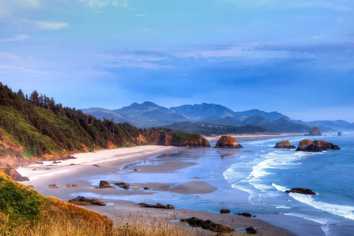 A sweeping view of Manzanita Beach, with gentle ocean waves lapping against the shore, distinctive rock formations, and mountains rising in the background. The tranquil scene invites exploration, beach walks, and family fun by the water.