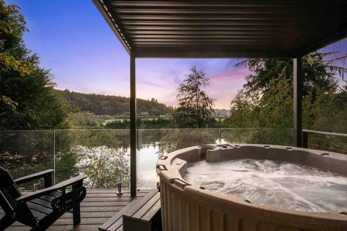 The image showcases an inviting deck nestled among towering trees, featuring a steaming hot tub with stunning views overlooking the calm Nehalem River. The serene setting is perfect for relaxation, whether soaking in the tub, watching a golden sunset over the water, or enjoying quiet conversation under the night sky.