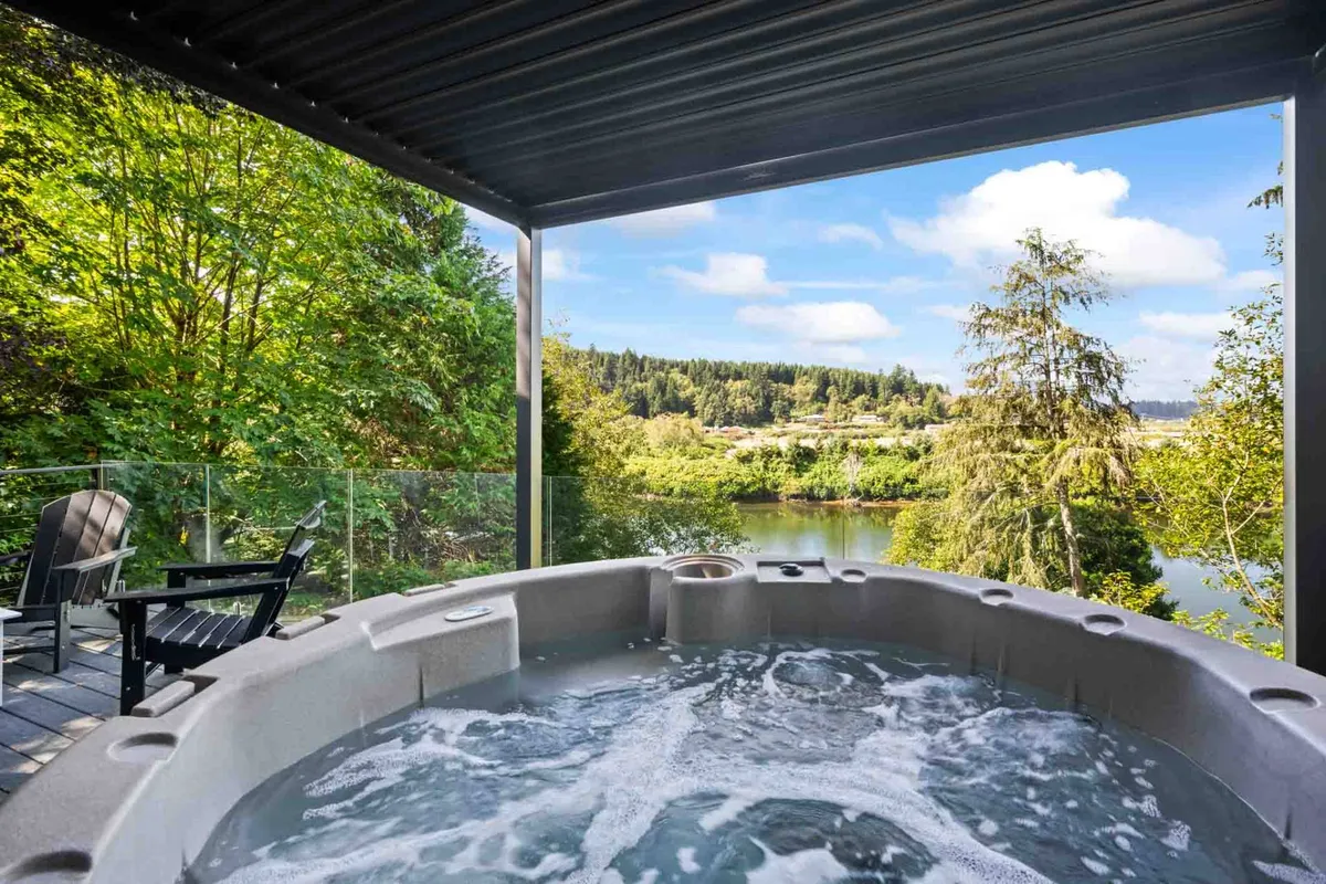 This image showcases an inviting hot tub on the deck overlooking the river, surrounded by lush greenery. The tranquil setting is perfect for relaxing in the evening, with clear views of the sky above for stargazing and space for friends or couples to unwind together.