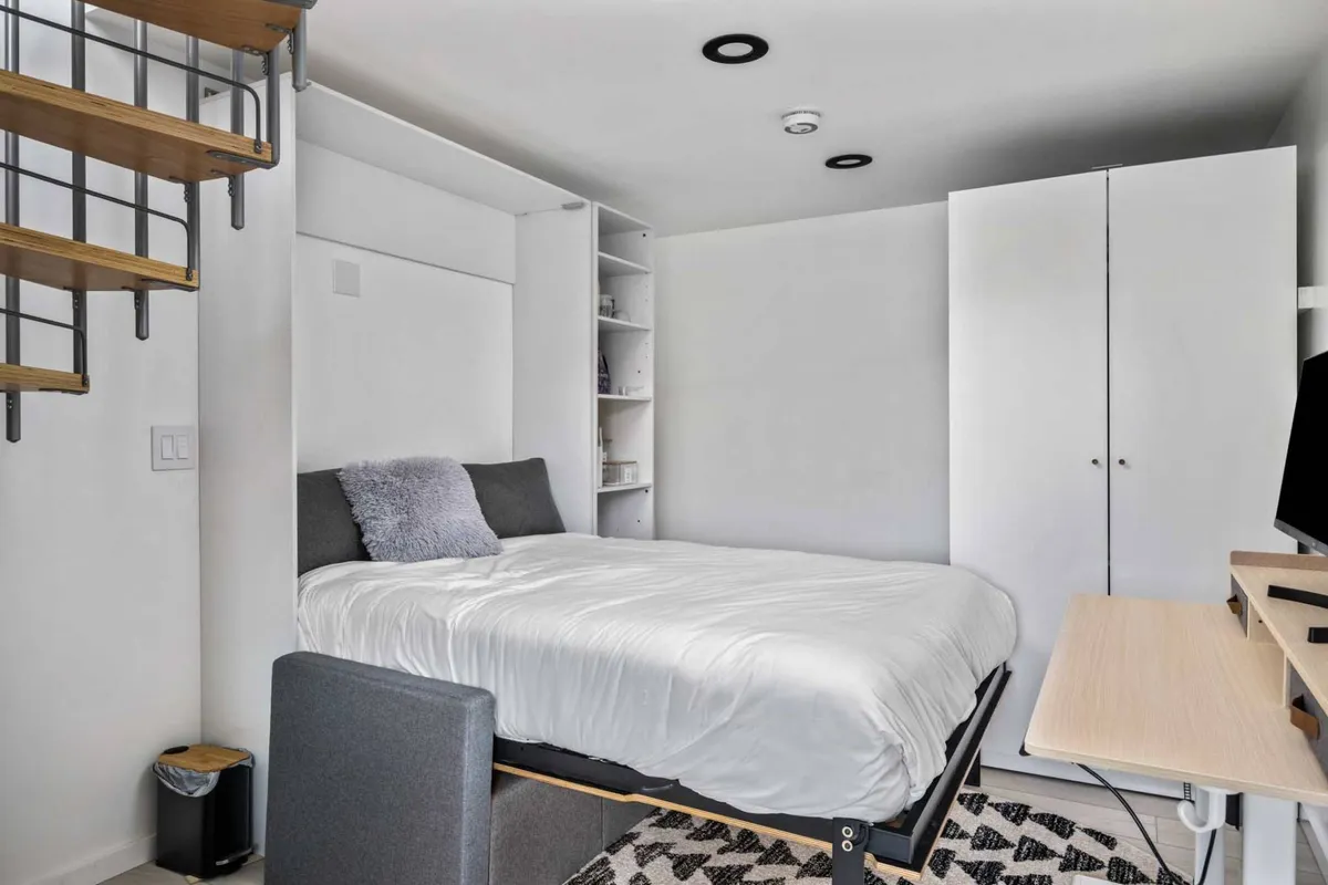 A cozy multipurpose room with a full bed nestled beside a modern staircase and a work desk, creating an adaptable space ideal for both rest and productivity. Soft lighting and minimal decor impart a sense of comfort and efficiency.